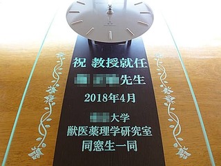 「祝教授就任」「○○先生」「日付」「○○大学○○研究室同窓生一同より」と、前面ガラスに彫刻した振り子型の電波式掛け時計(教授就任のお祝い品)