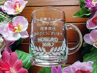 「社会医療法人社団○○、理事就任おめでとうございます。○○様、2018.7」を側面に彫刻した、理事就任祝いの贈り物用のガラス製マグカップ