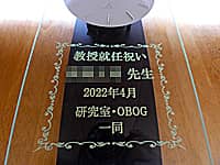 「教授就任祝い ○○先生 研究室・OBOG一同」を前面ガラスに彫刻した、教授就任祝い用の振り子時計
