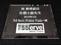 「祝 教授就任 ○○先生、○○一同」と「マーク」を彫刻した、教授就任祝い用のガラス盾